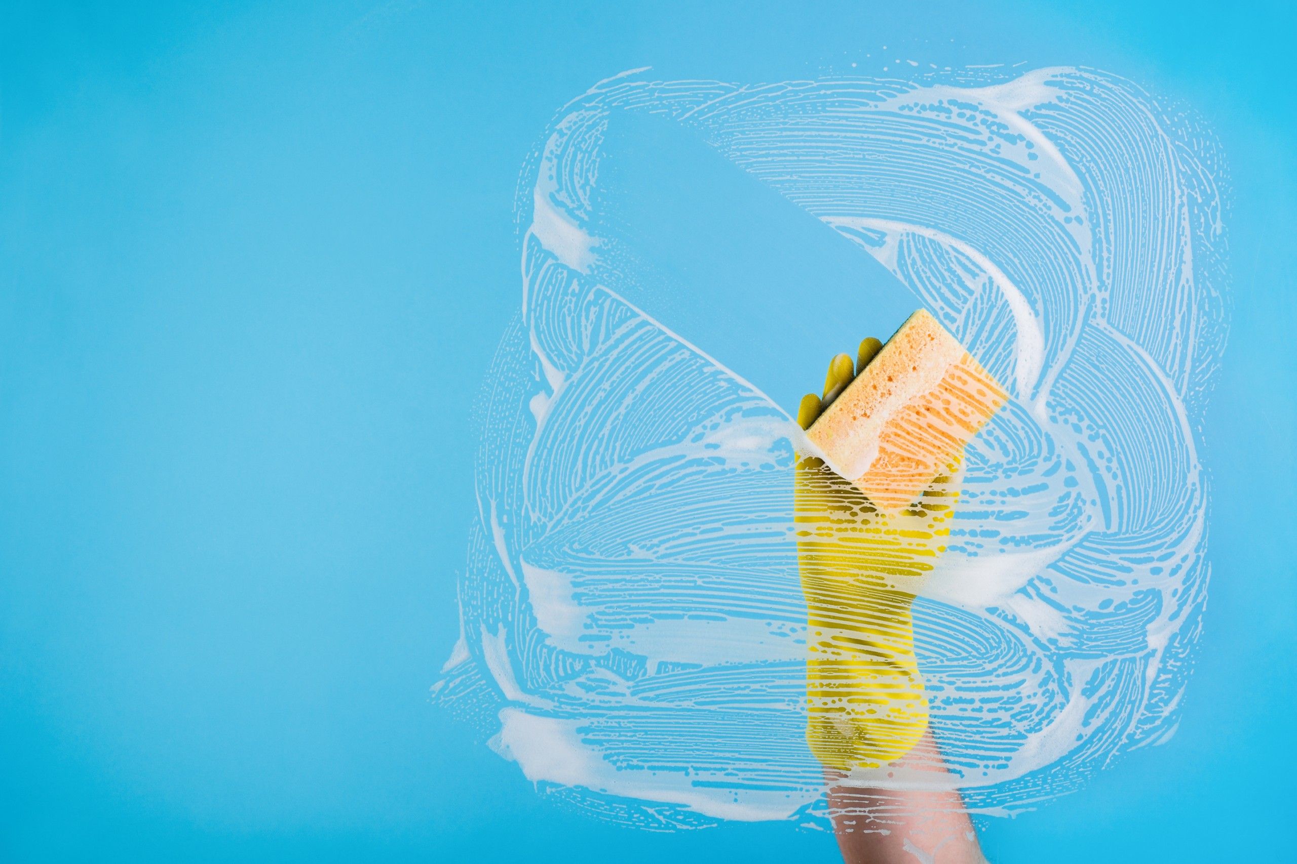 Gloved hand wiping soapy glass with a sponge against a bright blue background